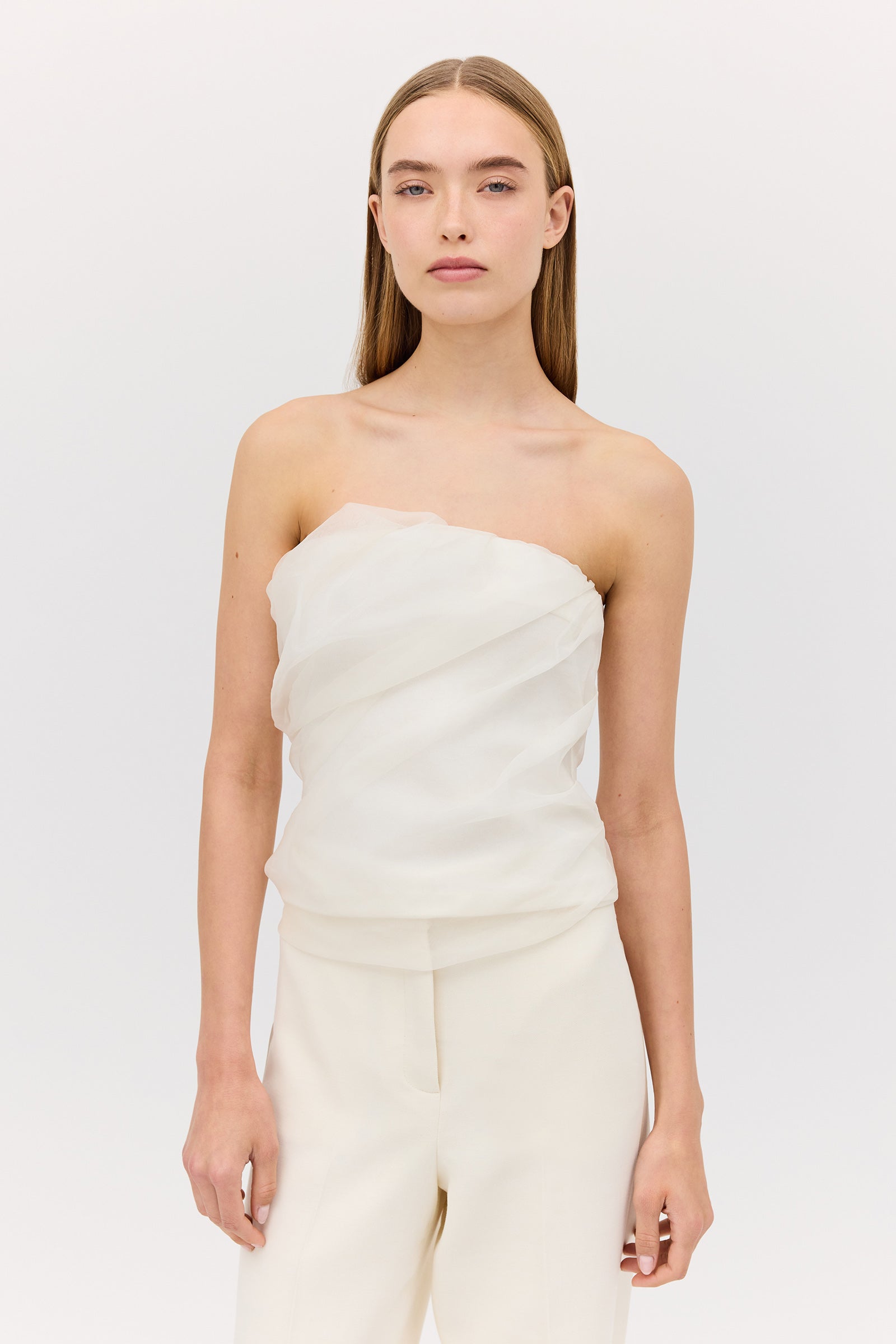 A woman wearing a cream-colored organza bustier top with a gathered detail, paired with white trousers.