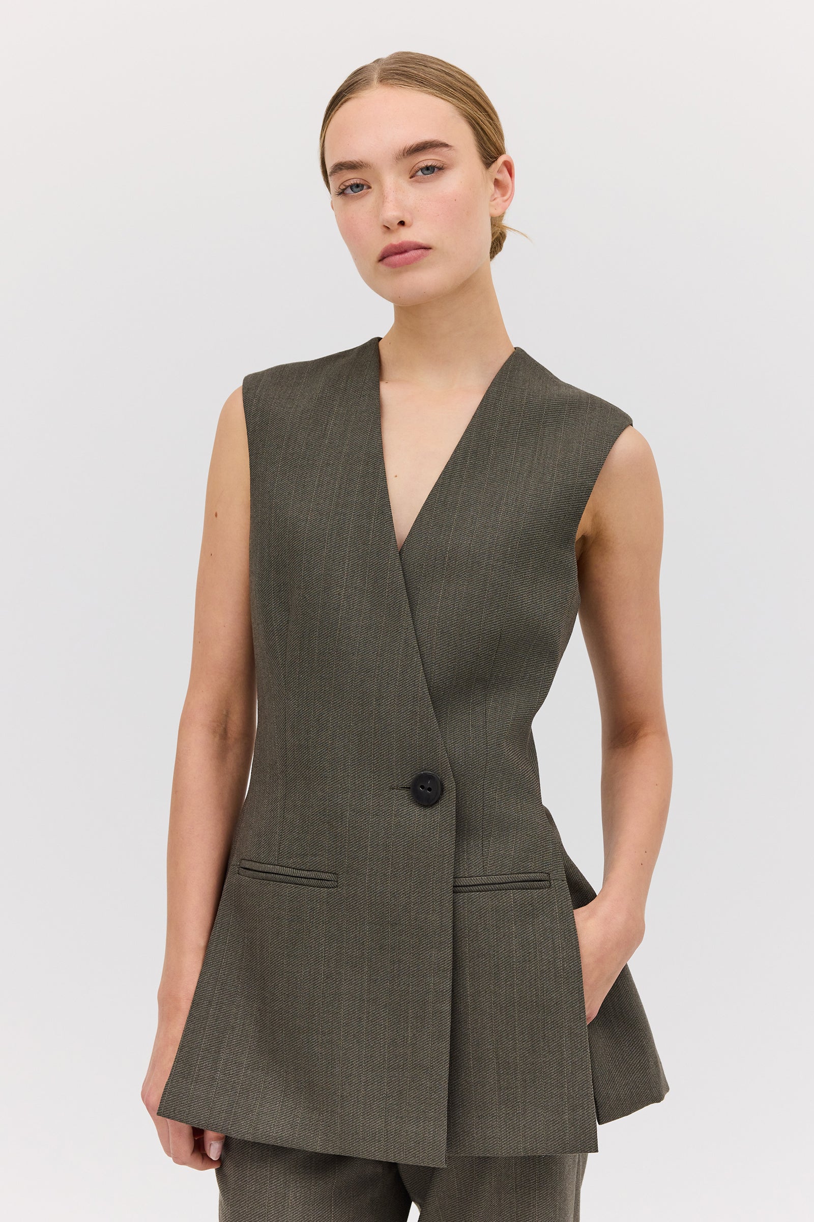 A woman modeling a sleeveless, single-breasted, mid-length Liquorice Wool Ellipsis Vest with functional front pockets, in a gray color.