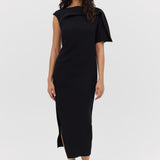 A woman standing in a studio setting, wearing a black midi-length dress with t-shirt sleeves and a boat neckline. The dress features fold and tuck detailing at the neck and shoulder.