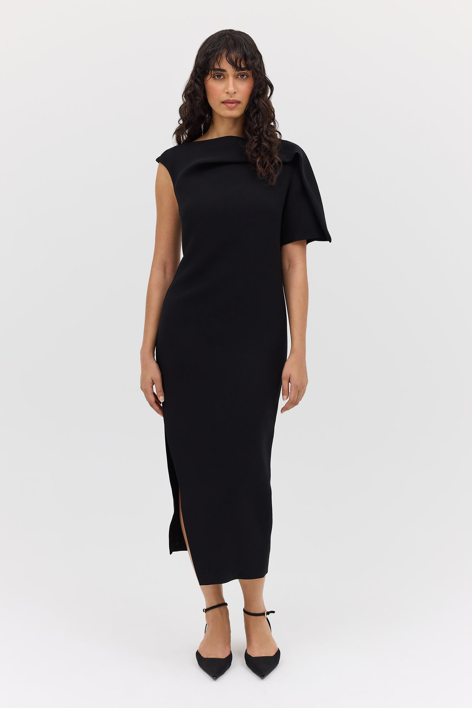 A woman standing in a studio setting, wearing a black midi-length dress with t-shirt sleeves and a boat neckline. The dress features fold and tuck detailing at the neck and shoulder.