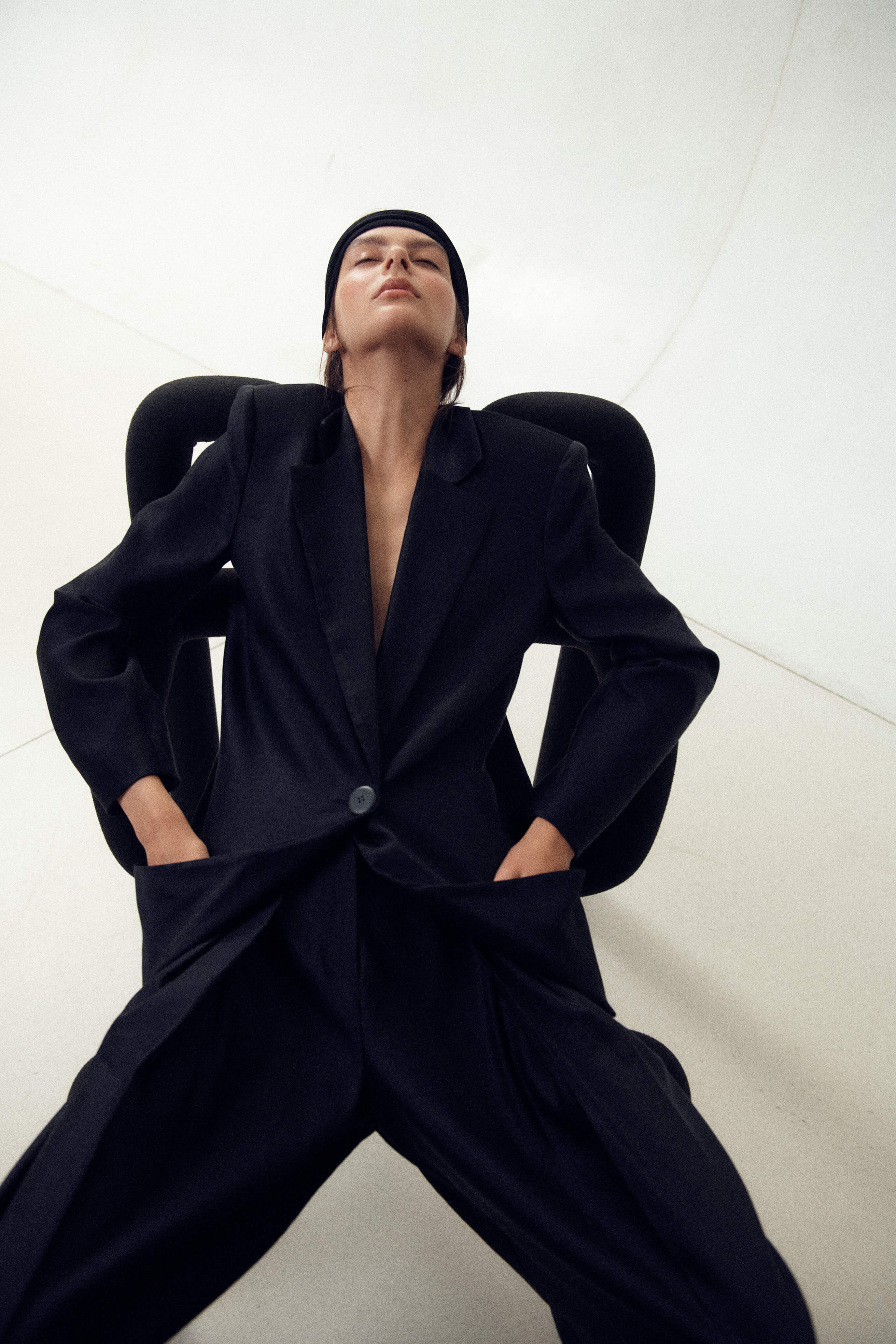 Model lays on chair with head band on and lack suit with hands in pockets.