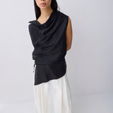 A model wearing a black sleeveless draped top with a drawstring waist and a flowing white skirt, standing against a plain light gray studio background.
