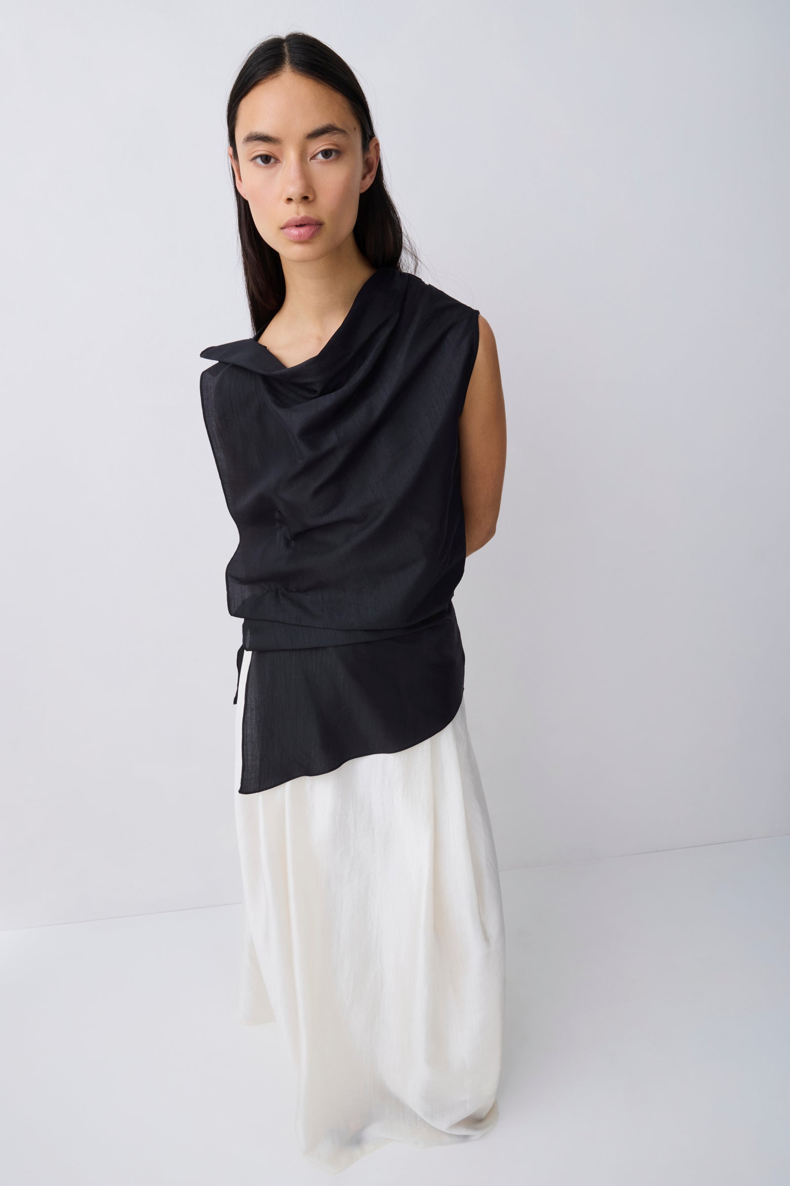 A model wearing a black sleeveless draped top with a drawstring waist and a flowing white skirt, standing against a plain light gray studio background.