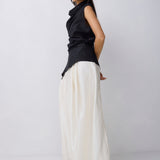 A model wearing a black sleeveless draped top and wide-leg white trousers, standing in profile against a light gray studio background.