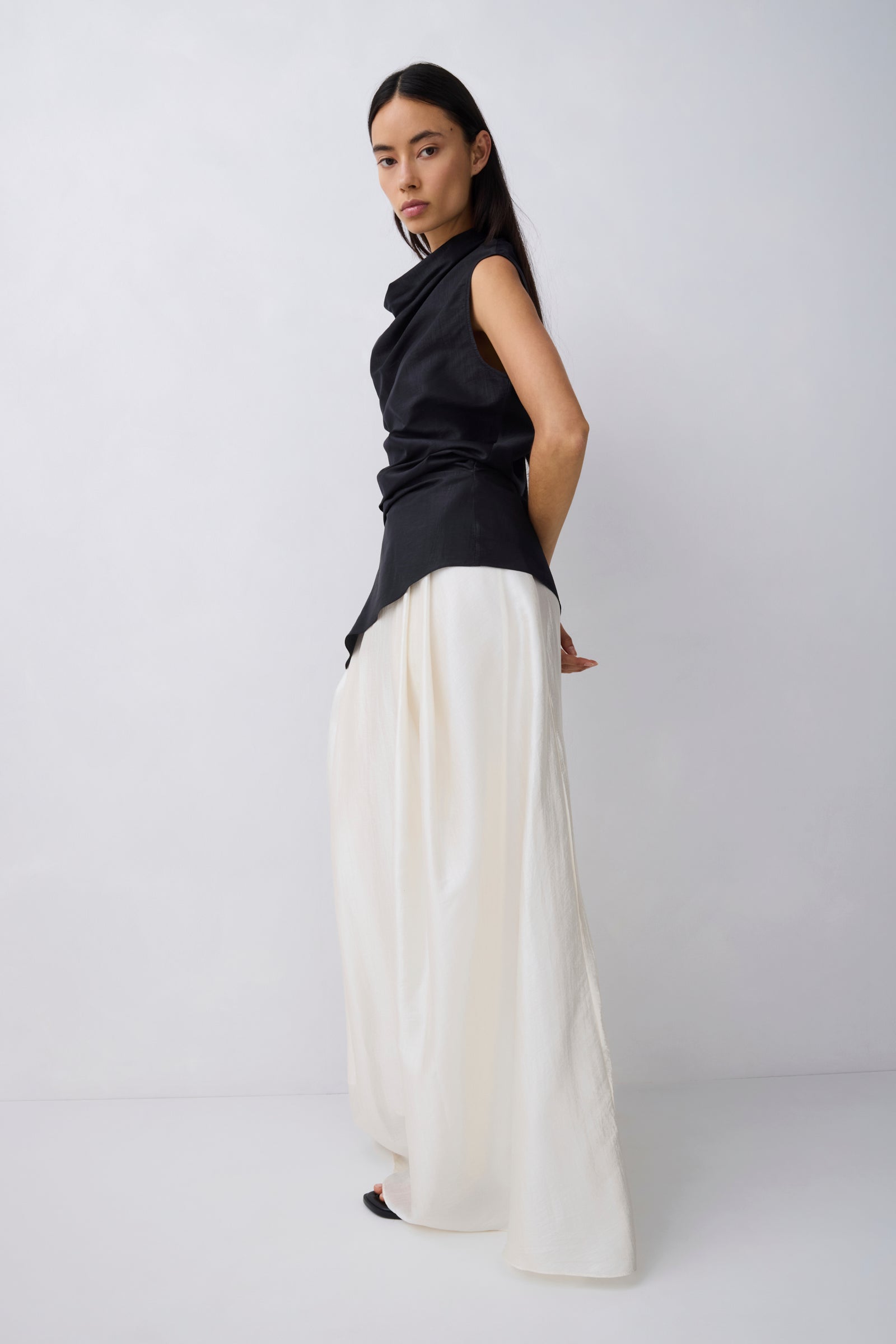 A model wearing a black sleeveless draped top and wide-leg white trousers, standing in profile against a light gray studio background.