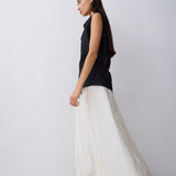 A woman in a sleeveless black top and a flowing white maxi skirt stands in profile against a plain light gray studio background.