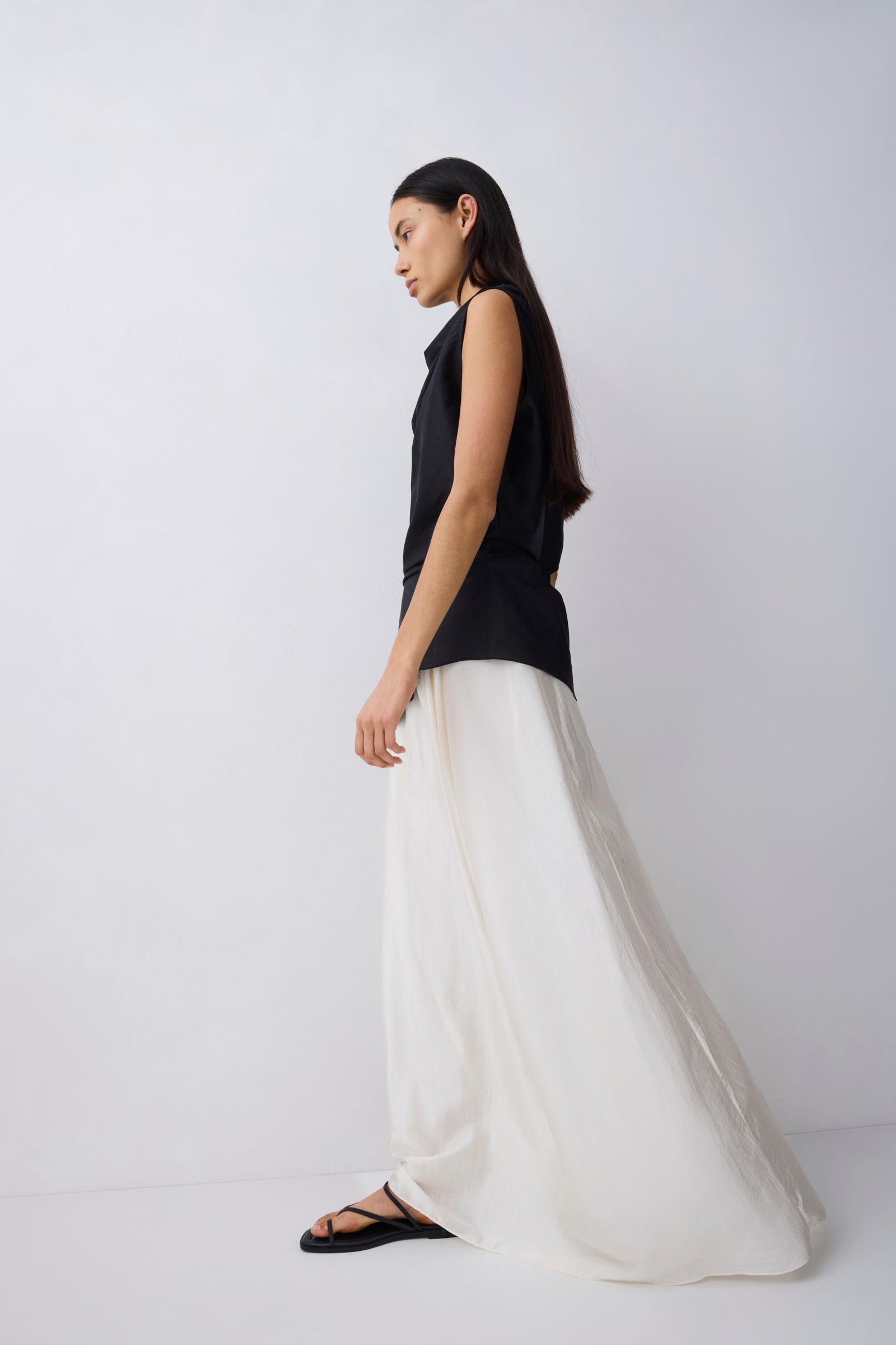 A woman in a sleeveless black top and a flowing white maxi skirt stands in profile against a plain light gray studio background.