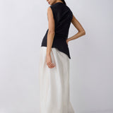 A model is shown from a side-back angle wearing a sleeveless black top and a long, flowing white skirt against a plain light gray studio background.