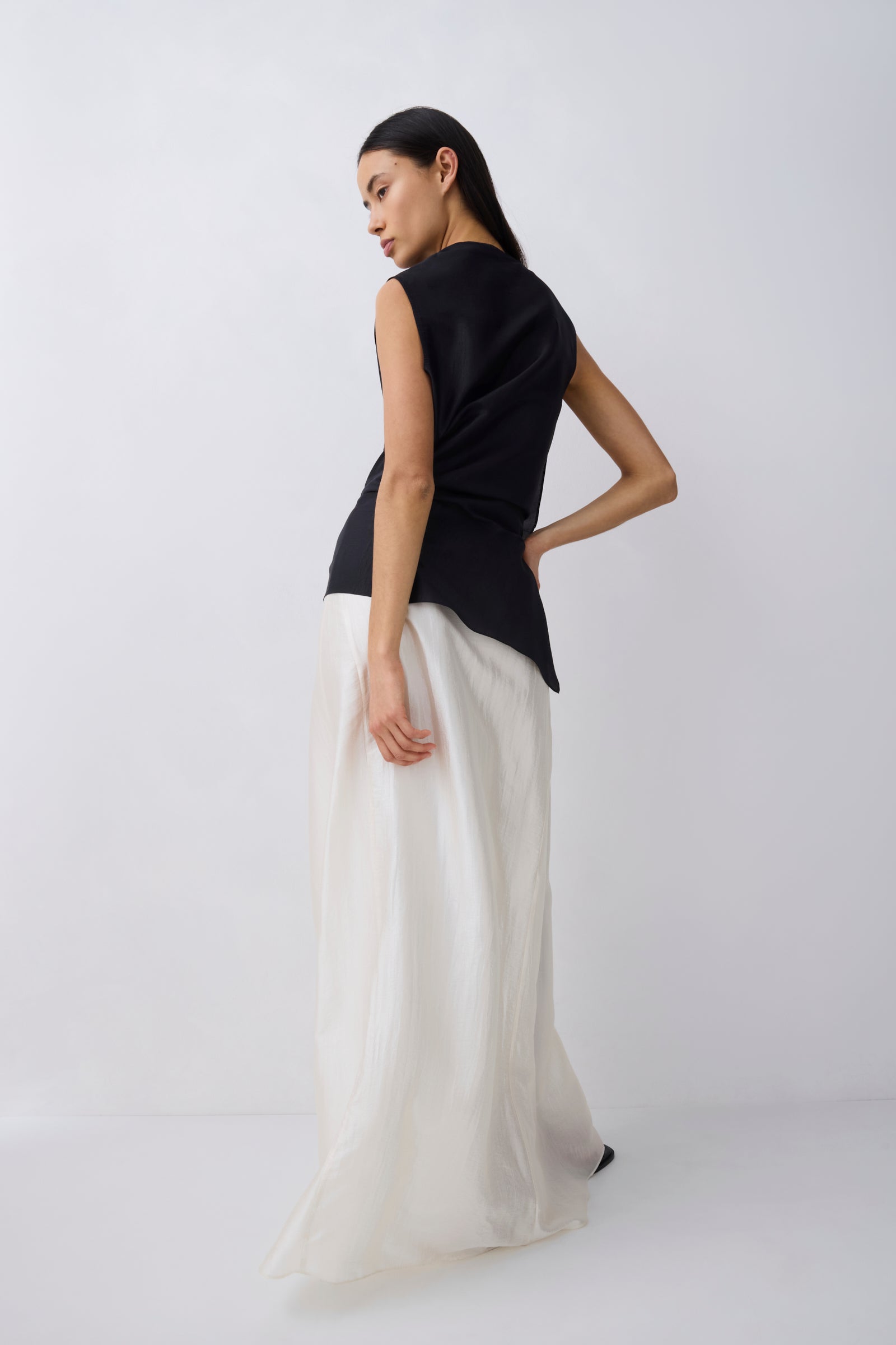 A model is shown from a side-back angle wearing a sleeveless black top and a long, flowing white skirt against a plain light gray studio background.