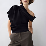A model wearing a black draped off-the-shoulder top and dark olive trousers, standing against a light gray studio background.