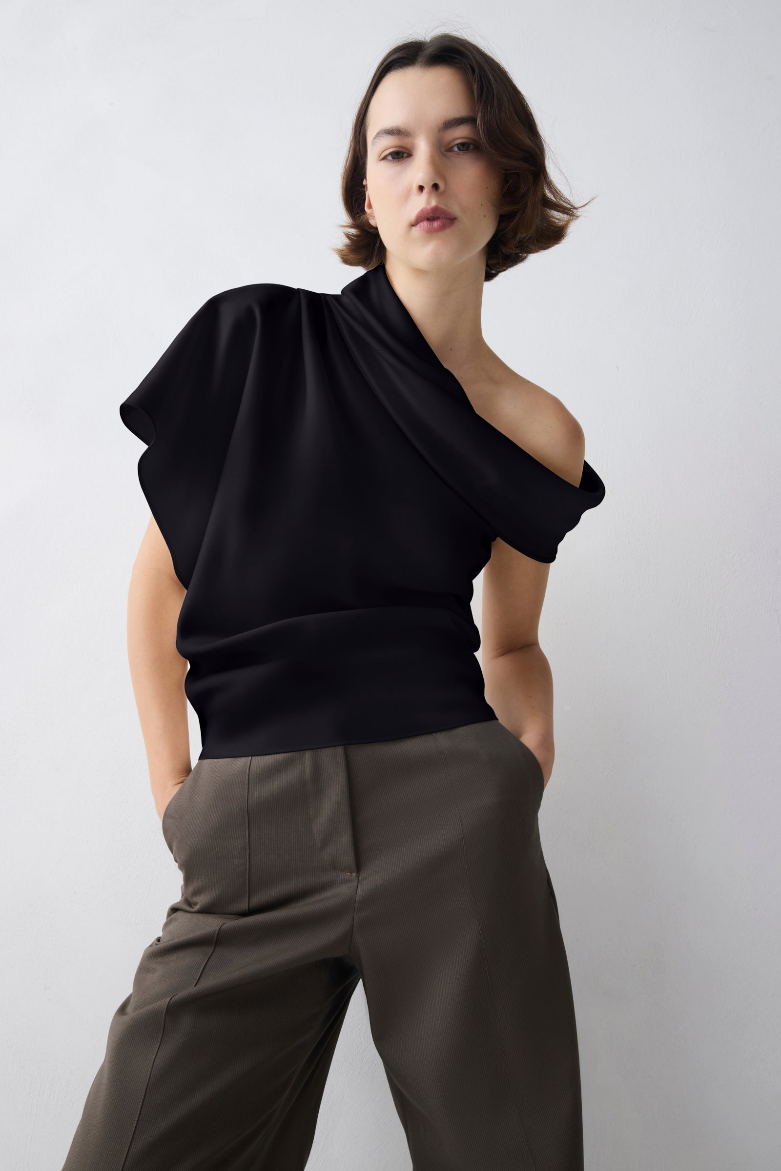 A model wearing a black draped off-the-shoulder top and dark olive trousers, standing against a light gray studio background.
