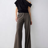 A model stands against a light gray studio background wearing a black one-shoulder draped top and high-waisted, wide-leg taupe-brown trousers.