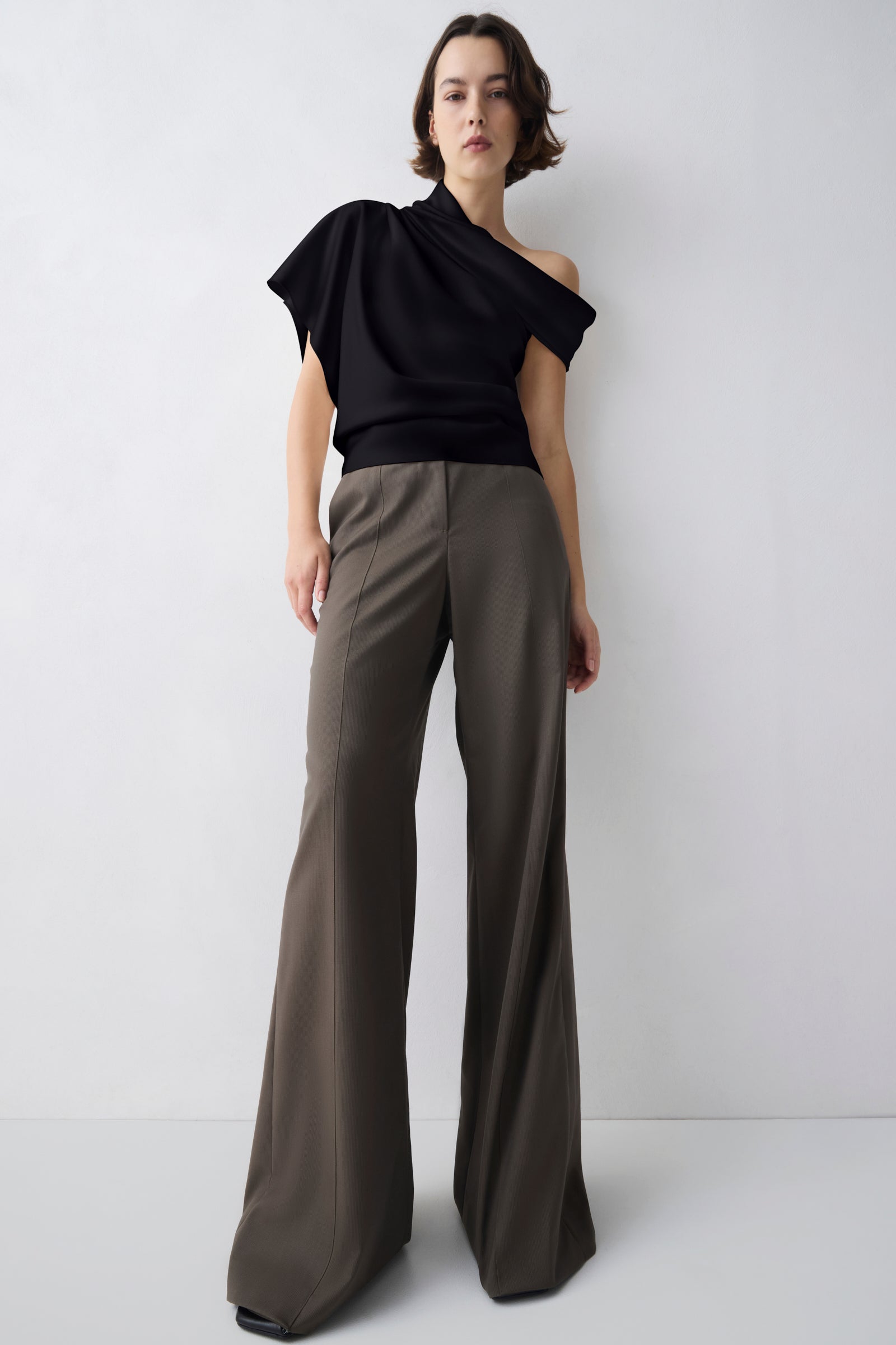 A model stands against a light gray studio background wearing a black one-shoulder draped top and high-waisted, wide-leg taupe-brown trousers.