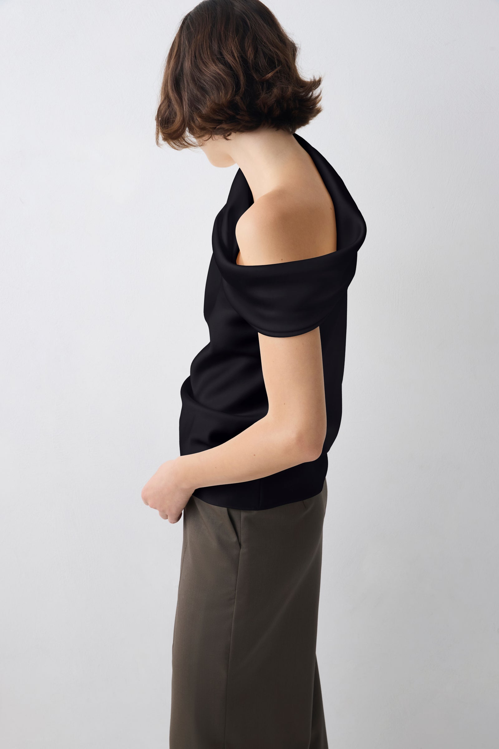 A model shown from the side and back wearing a black off-the-shoulder knit top and dark olive trousers, set against a plain light gray studio background.