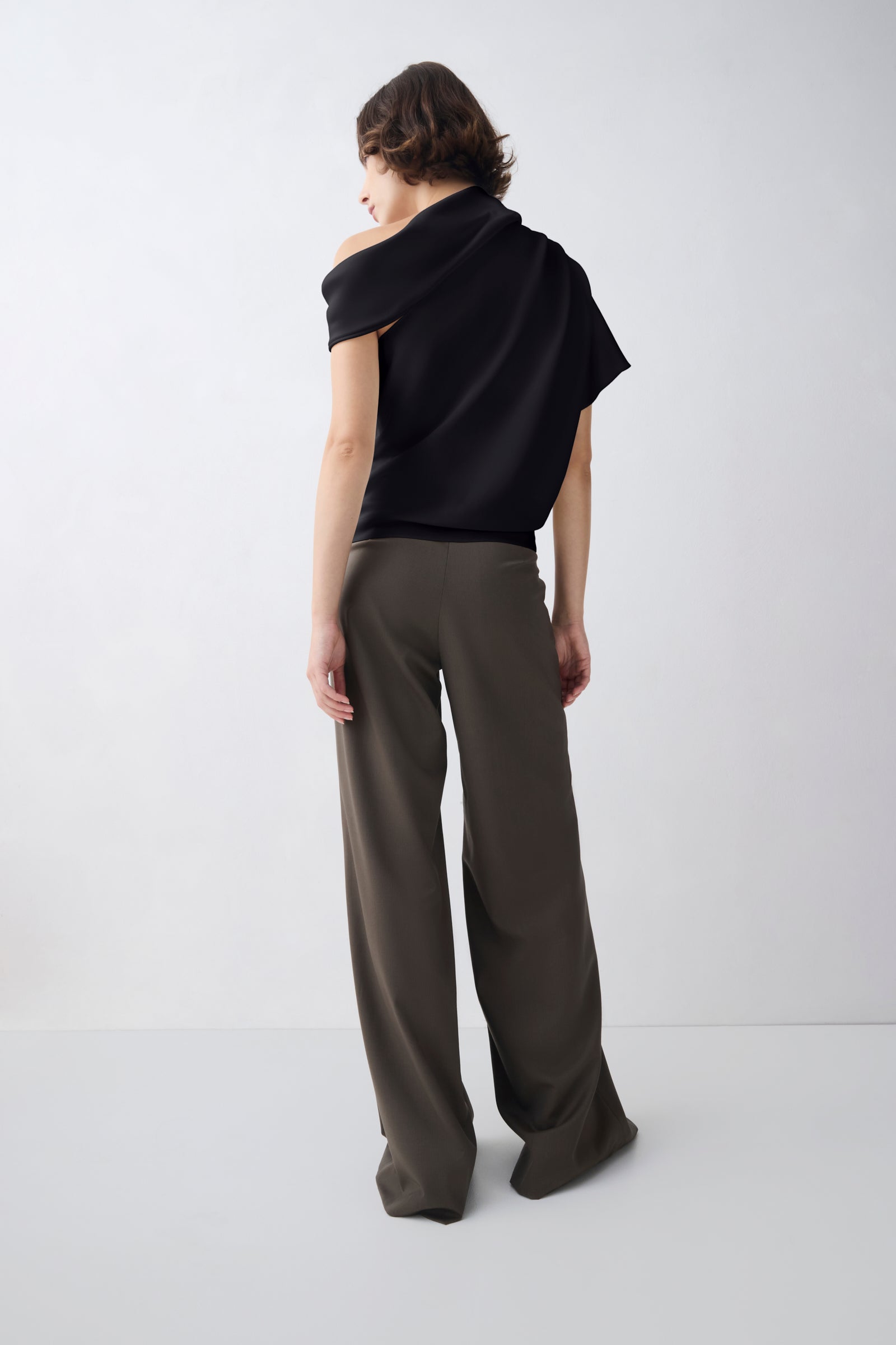 A model shown from the back wearing a black draped short-sleeve top and wide-leg olive brown trousers, standing against a light gray studio background.