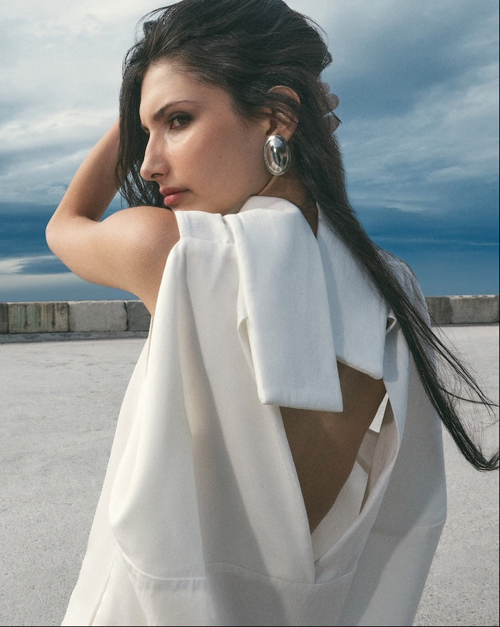 A woman with long dark hair stands on a rooftop wearing a white draped wrap dress with a large bow on the shoulder and an open back, set against a cloudy blue sky.
