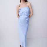 A woman modeling a light blue draped spaghetti-strap maxi dress with black flat shoes, standing against a plain white studio background.