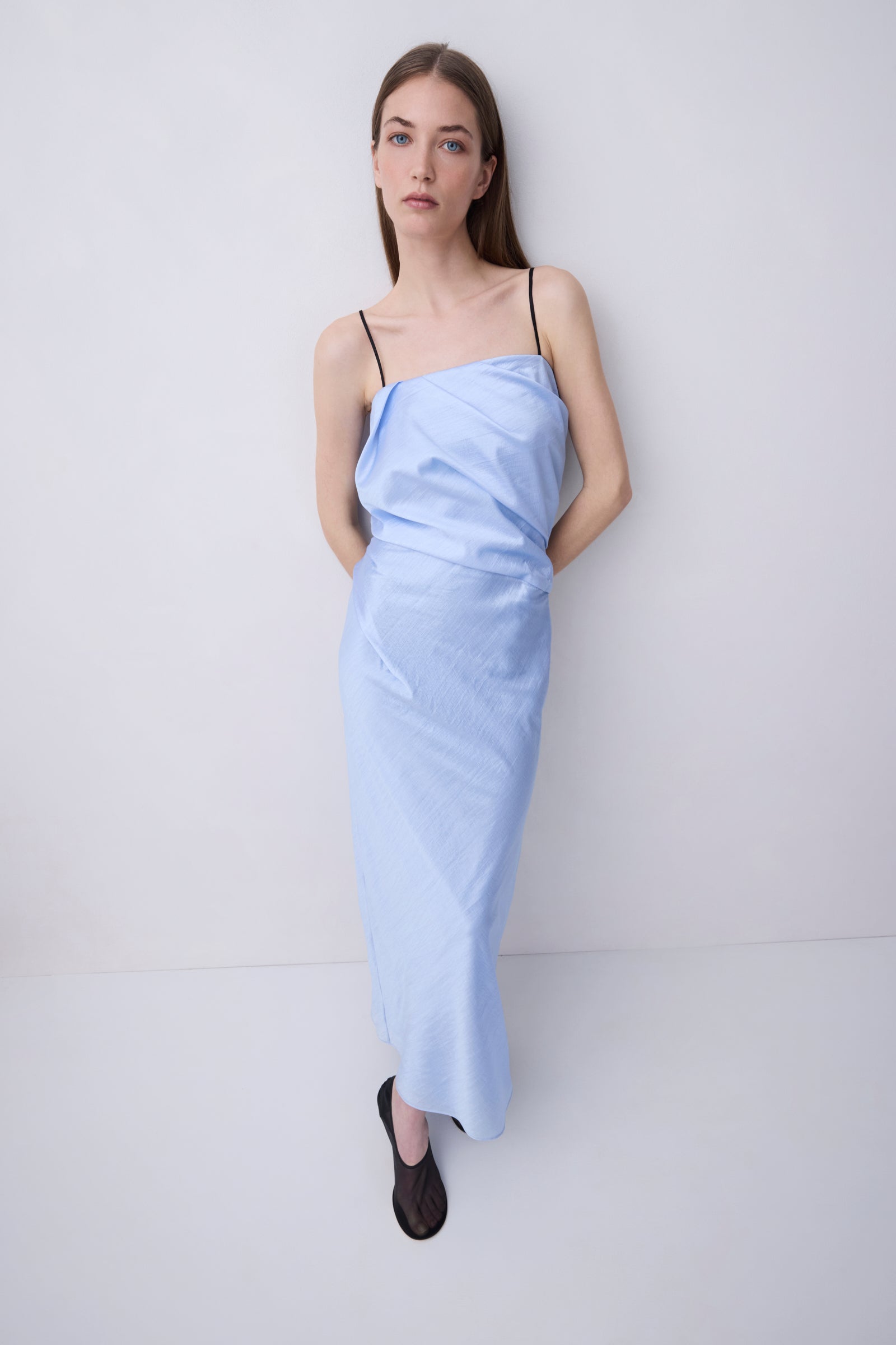 A woman modeling a light blue draped spaghetti-strap maxi dress with black flat shoes, standing against a plain white studio background.
