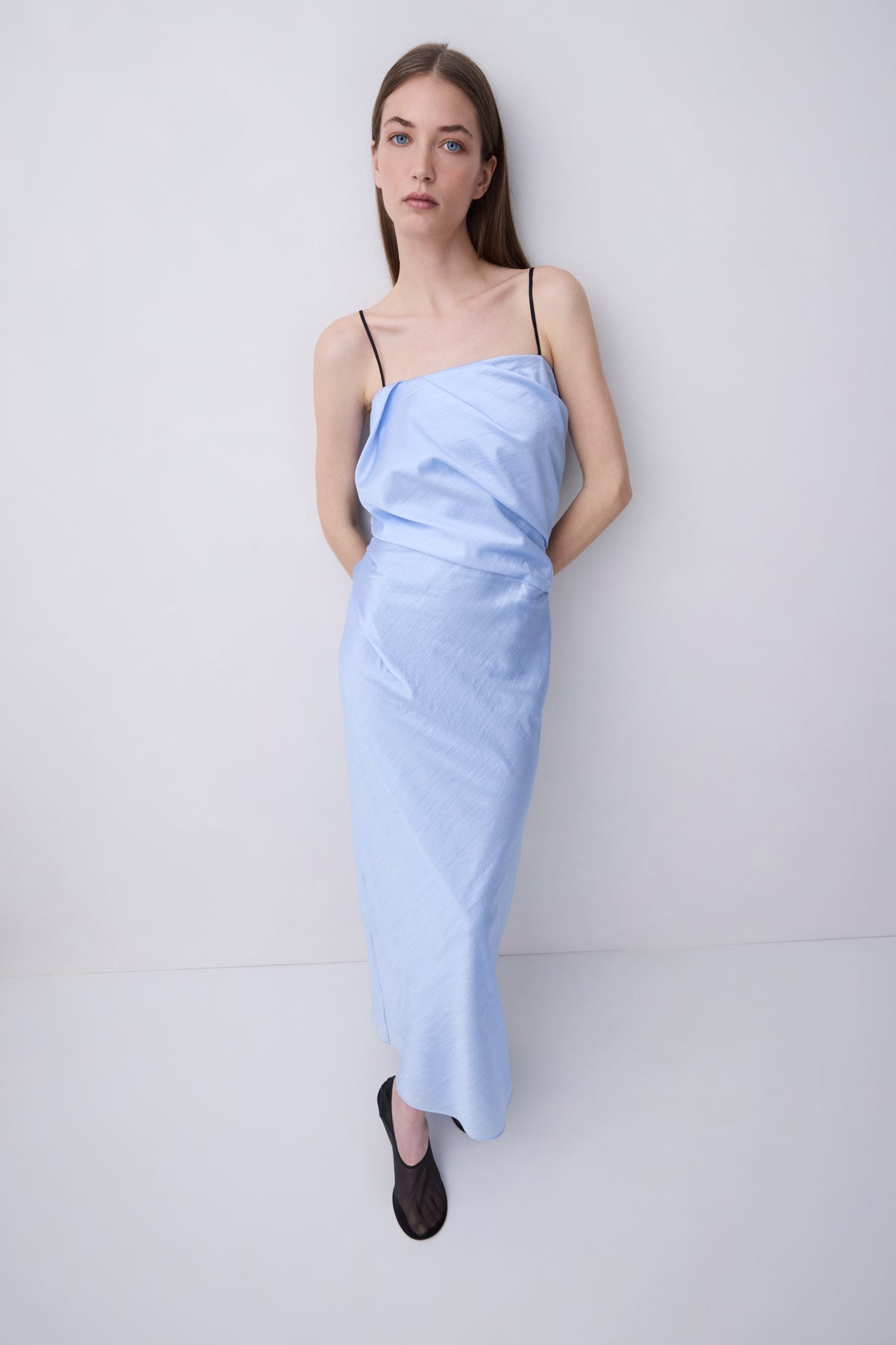 A woman modeling a light blue draped spaghetti-strap maxi dress with black flat shoes, standing against a plain white studio background.