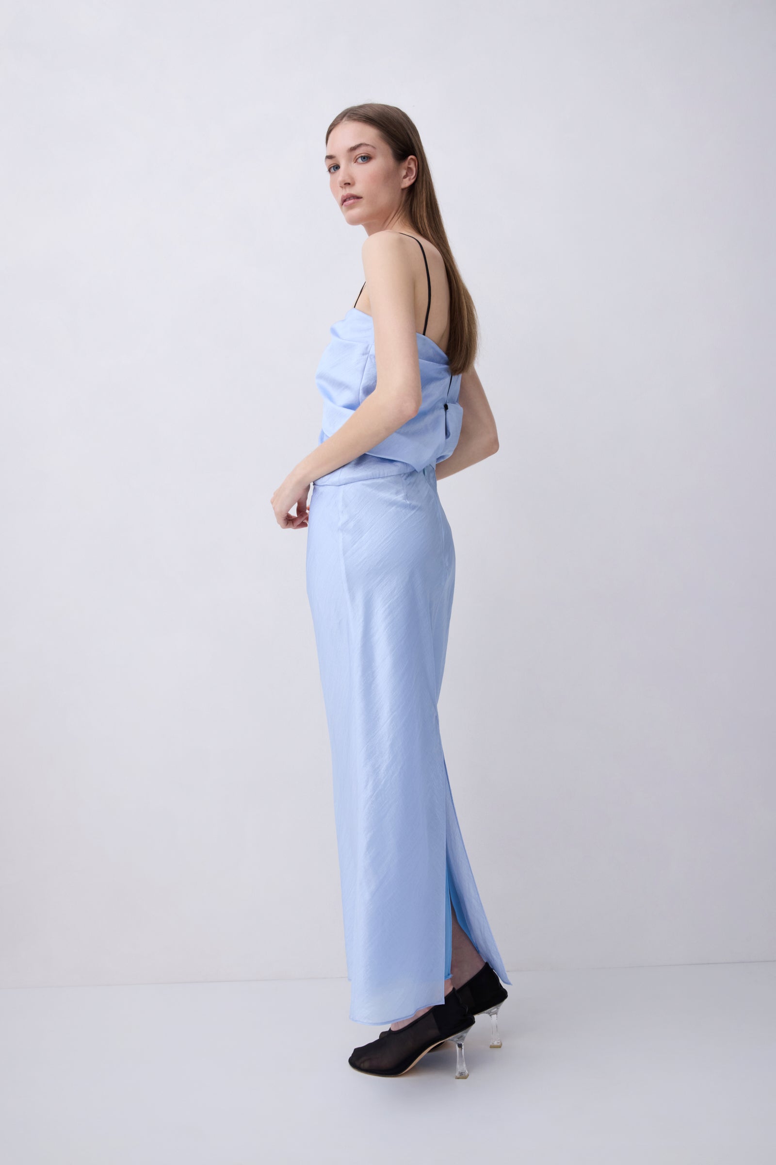 A woman modeling a light blue strapless maxi dress with a side slit, paired with black heels, standing against a plain light gray studio background.