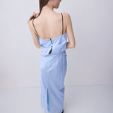 A woman viewed from behind wearing a light blue maxi dress with thin black spaghetti straps, a draped open back, and a side slit, set against a plain light gray studio background.