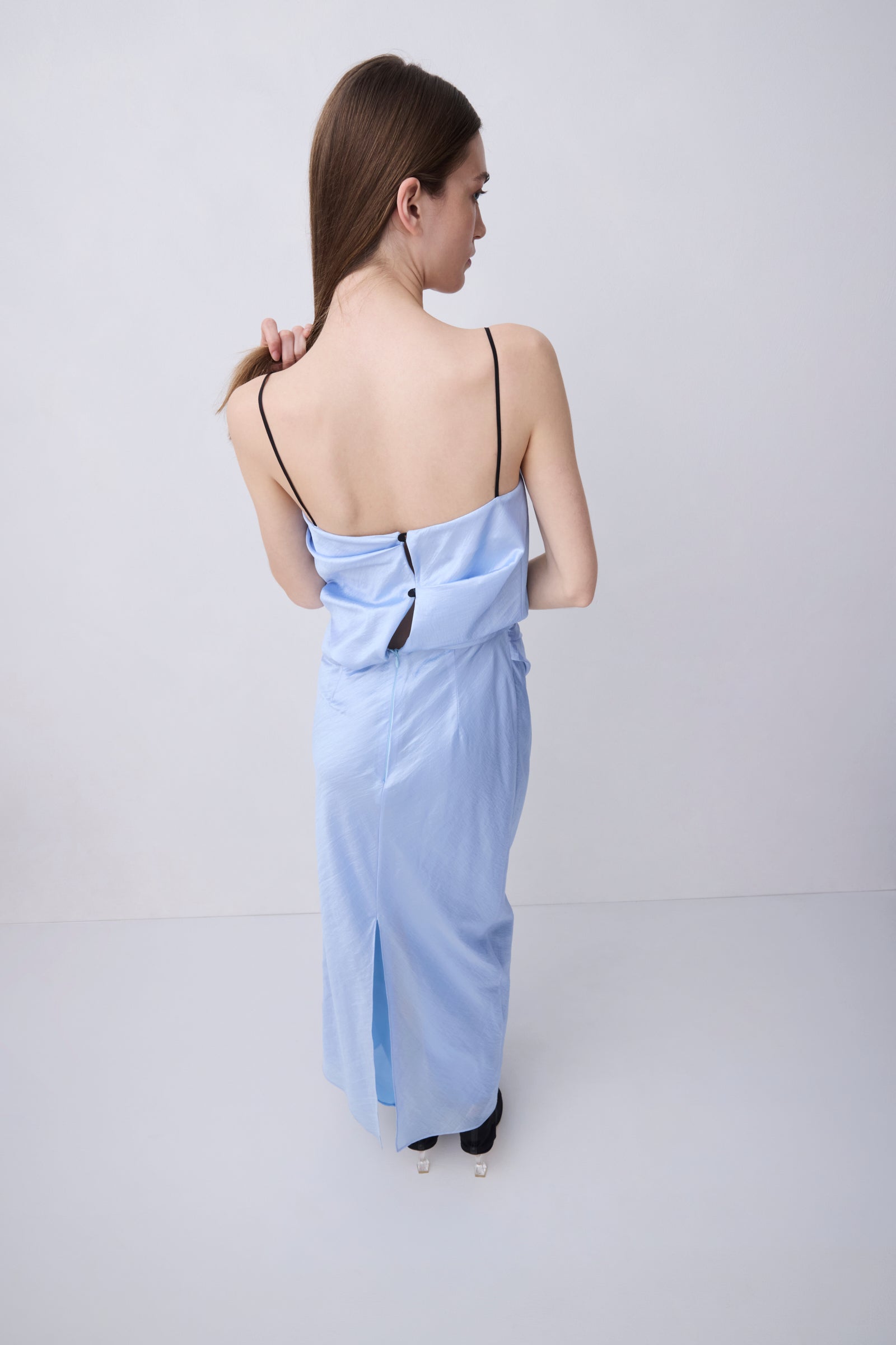 A woman viewed from behind wearing a light blue maxi dress with thin black spaghetti straps, a draped open back, and a side slit, set against a plain light gray studio background.