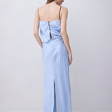 A woman stands with her back to the camera wearing a light blue maxi dress with thin black spaghetti straps, a wrap-style back with a central cutout, and a front slit, paired with black high heels against a plain light gray studio background.