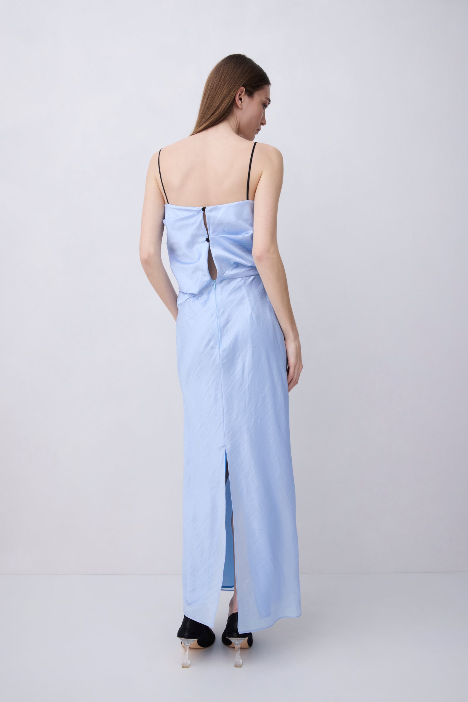 A woman stands with her back to the camera wearing a light blue maxi dress with thin black spaghetti straps, a wrap-style back with a central cutout, and a front slit, paired with black high heels against a plain light gray studio background.