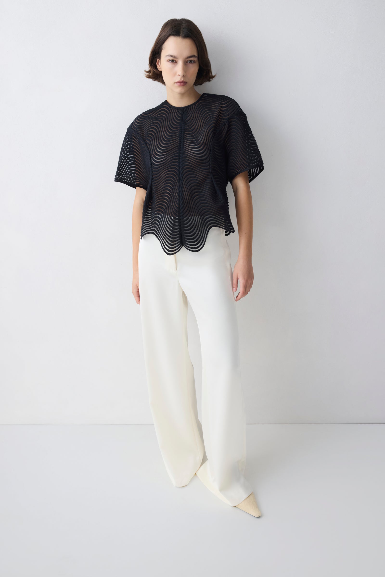Person wearing a black textured top and white pants on a white background