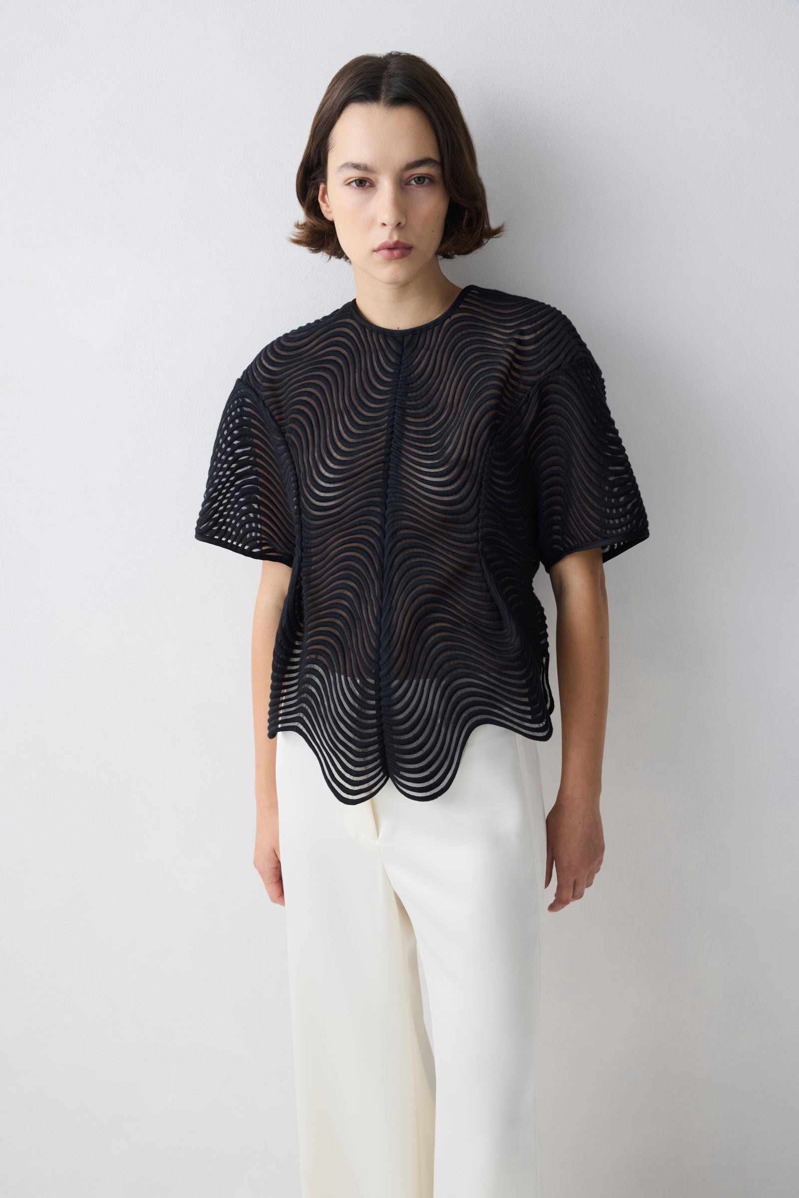 Person wearing a black, textured top with white pants on a plain background