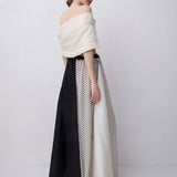 A model is shown from the back wearing a unique off-the-shoulder maxi dress with a textured cream top and a skirt featuring panels of solid black, white, and black-and-white polka dot pattern.