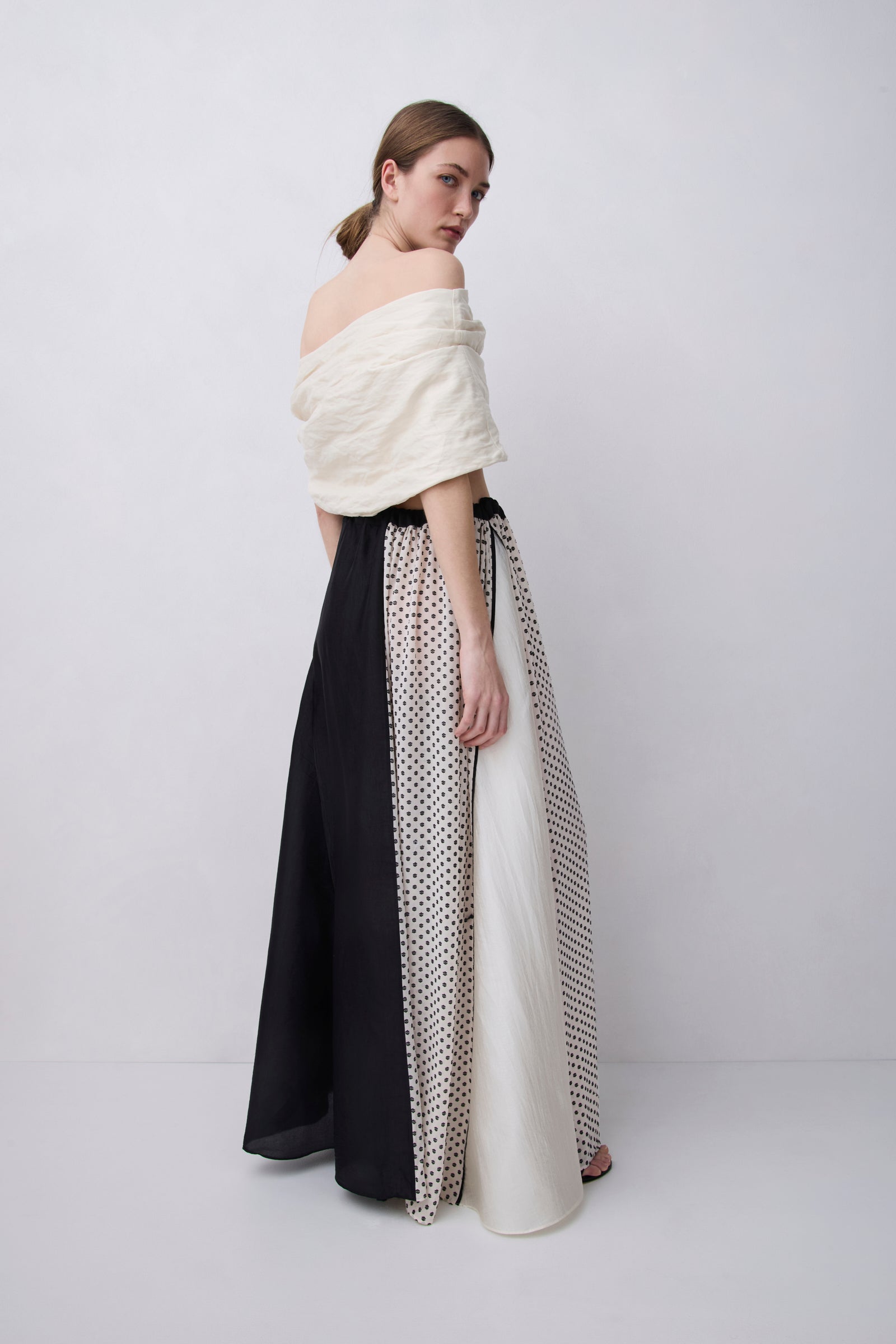 A model is shown from the back wearing a unique off-the-shoulder maxi dress with a textured cream top and a skirt featuring panels of solid black, white, and black-and-white polka dot pattern.