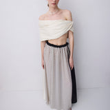 A model stands against a light gray studio background wearing a draped off-the-shoulder cream top and a long skirt with a black and white polka dot pattern and a black side panel.