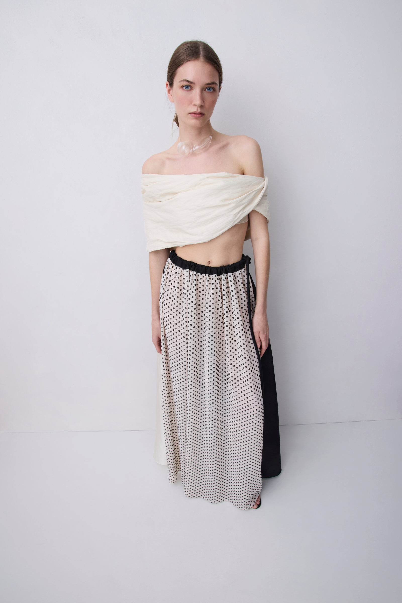 A model stands against a light gray studio background wearing a draped off-the-shoulder cream top and a long skirt with a black and white polka dot pattern and a black side panel.