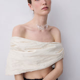 A model wearing a cream-colored crinkled fabric wrap or shawl draped off her shoulders, paired with a black and white polka dot skirt, against a plain light gray background.