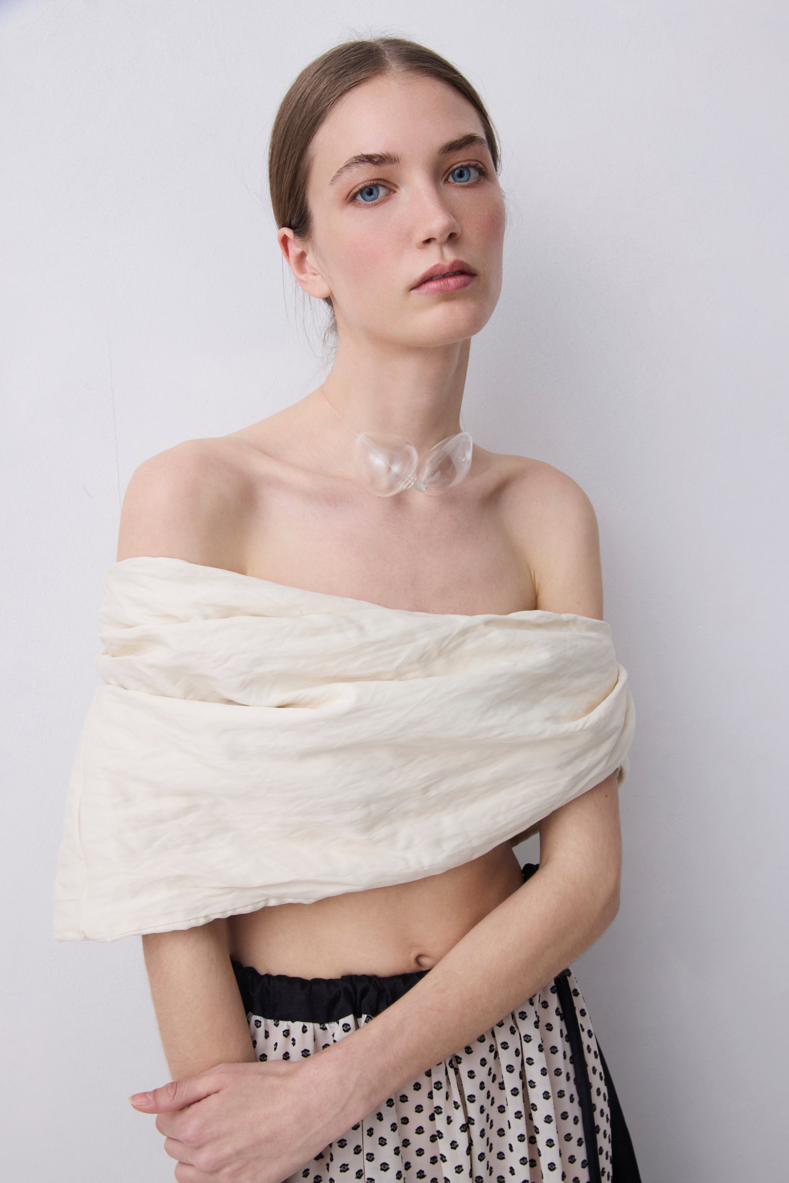 A model wearing a cream-colored crinkled fabric wrap or shawl draped off her shoulders, paired with a black and white polka dot skirt, against a plain light gray background.