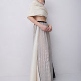 A model stands in a studio wearing a long cream and black polka-dot skirt with a sheer side panel and a cropped cream top draped over her shoulders.
