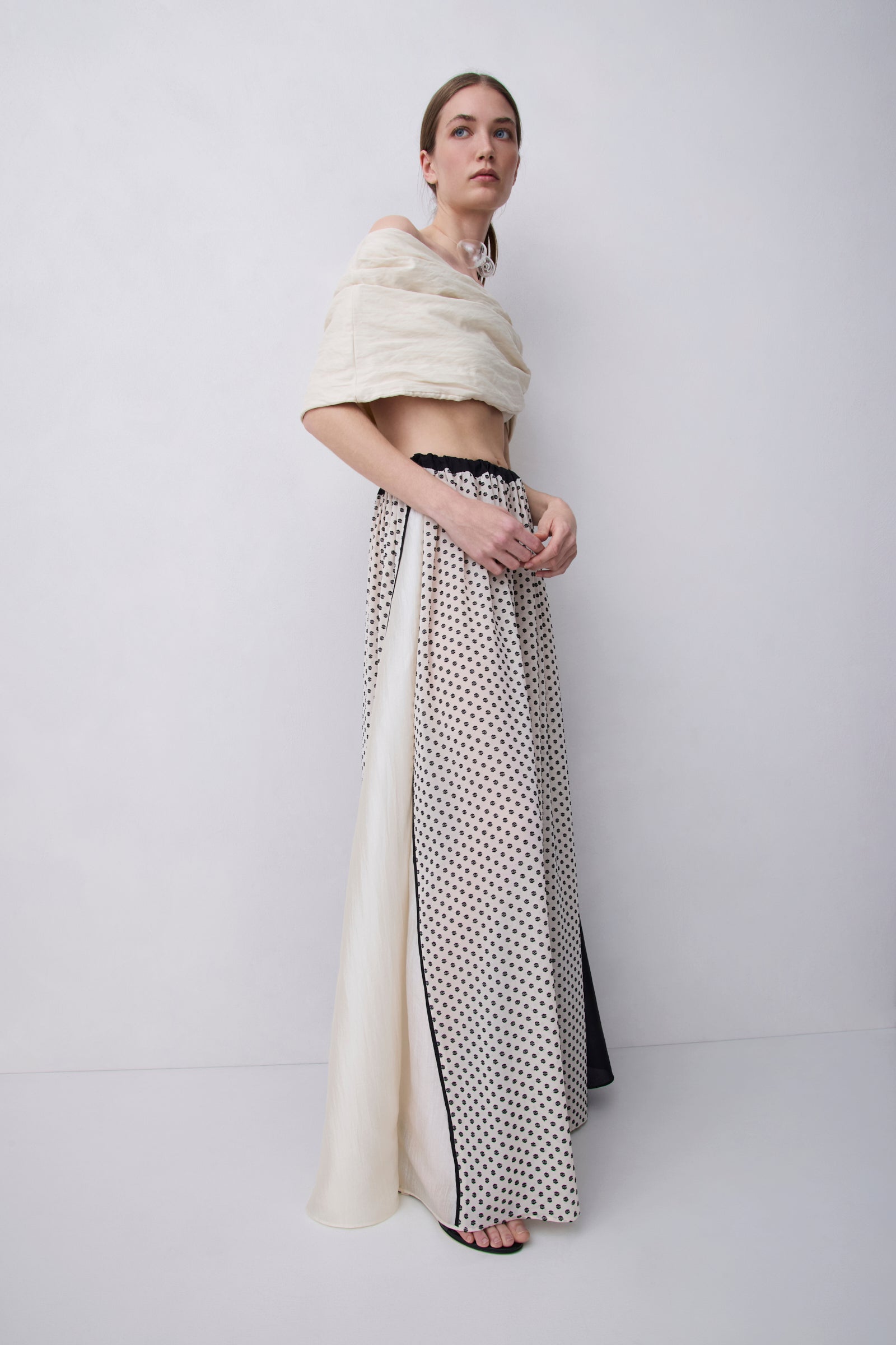 A model stands in a studio wearing a long cream and black polka-dot skirt with a sheer side panel and a cropped cream top draped over her shoulders.