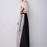 A model wearing a long, off-the-shoulder dress with a black and white polka dot pattern on one side and solid black on the other, paired with a cream draped wrap top, set against a plain light gray studio background.