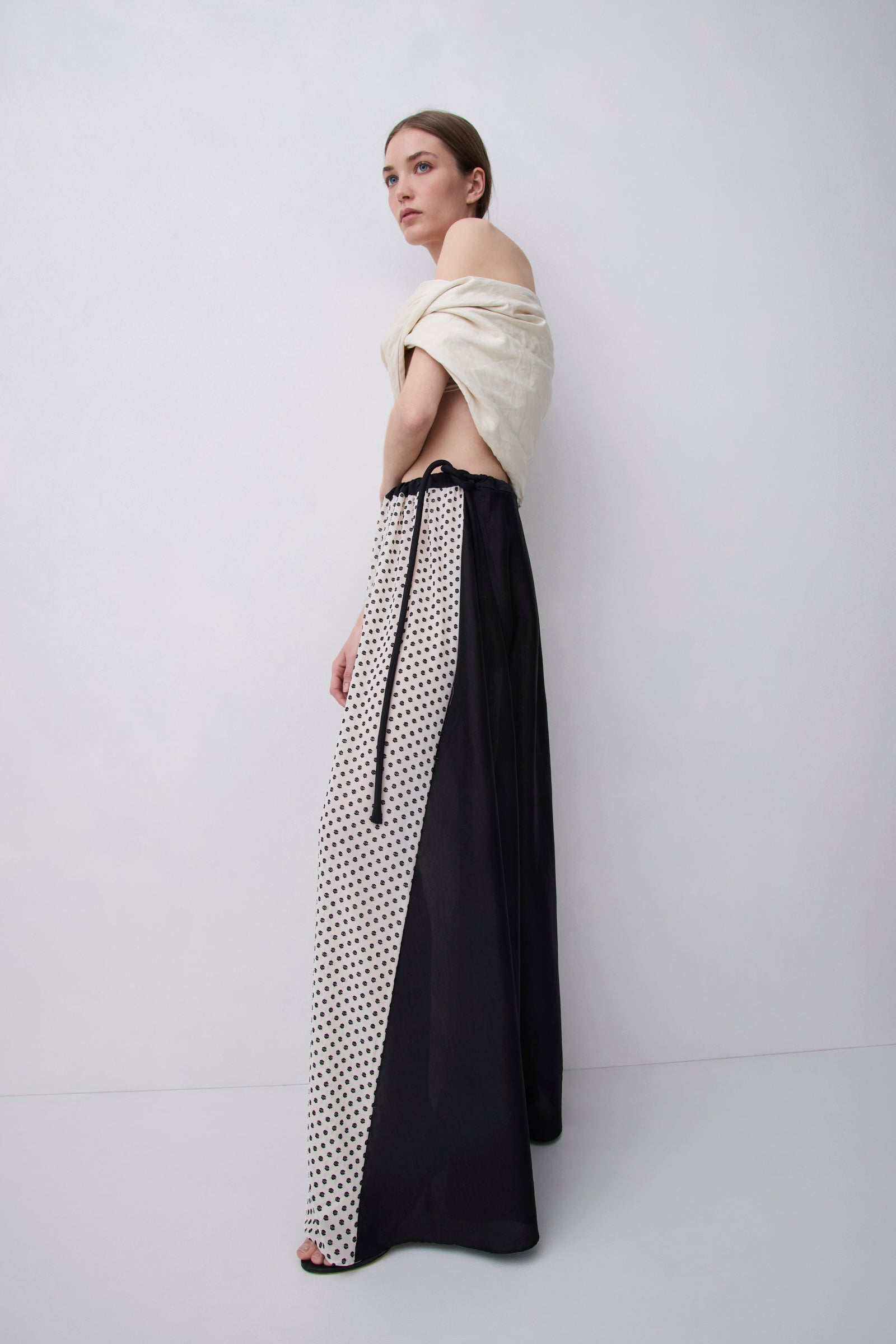 A model wearing a long, off-the-shoulder dress with a black and white polka dot pattern on one side and solid black on the other, paired with a cream draped wrap top, set against a plain light gray studio background.