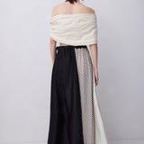 A model shown from the back wearing a unique off-the-shoulder maxi dress with a cream textured top, a black skirt panel, and a cream panel with black polka dots.