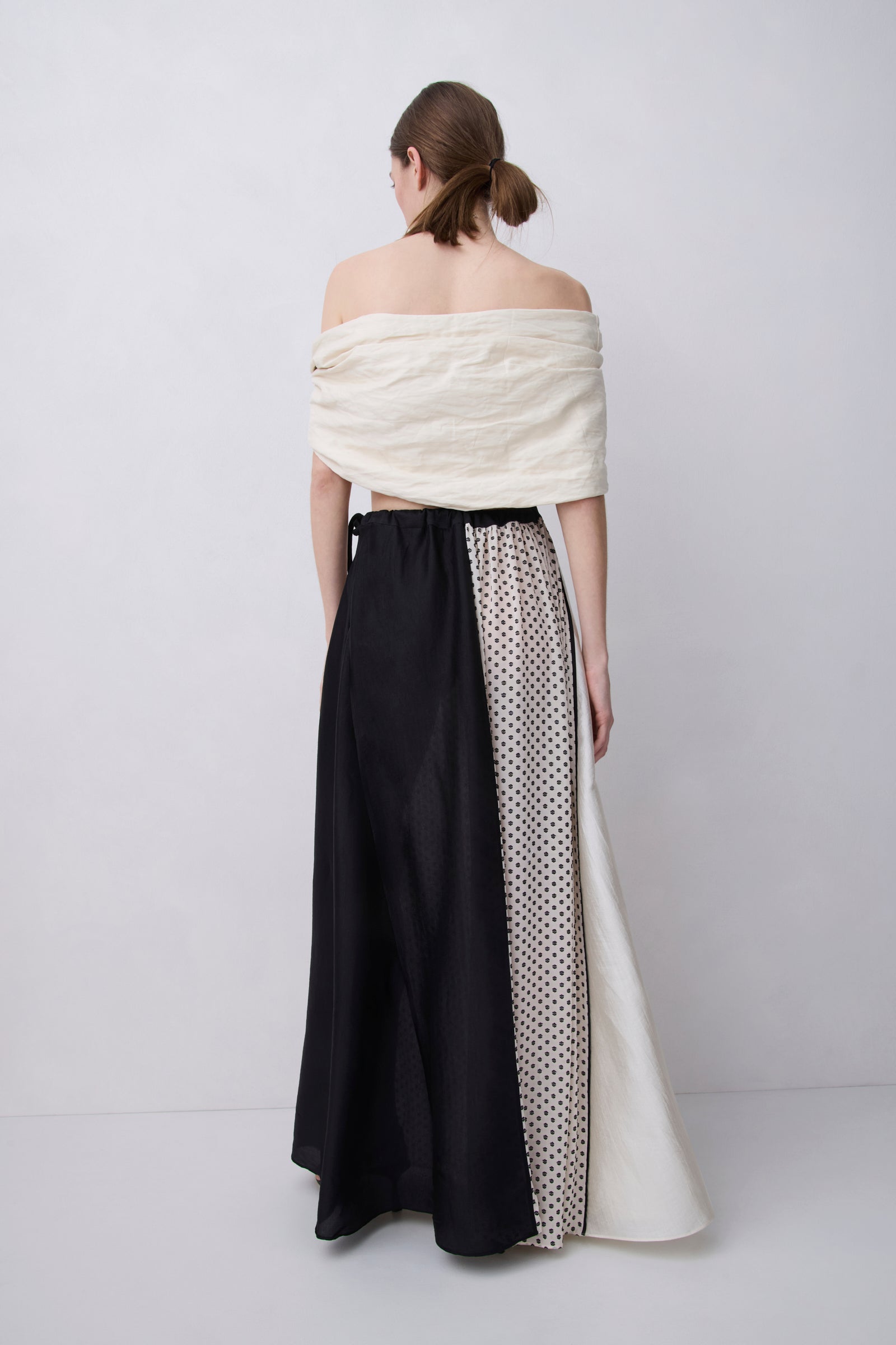 A model shown from the back wearing a unique off-the-shoulder maxi dress with a cream textured top, a black skirt panel, and a cream panel with black polka dots.
