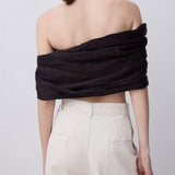 A woman shown from the back wearing a black off-the-shoulder ruched crop top and cream-colored wide-leg trousers, set against a plain light gray studio background.