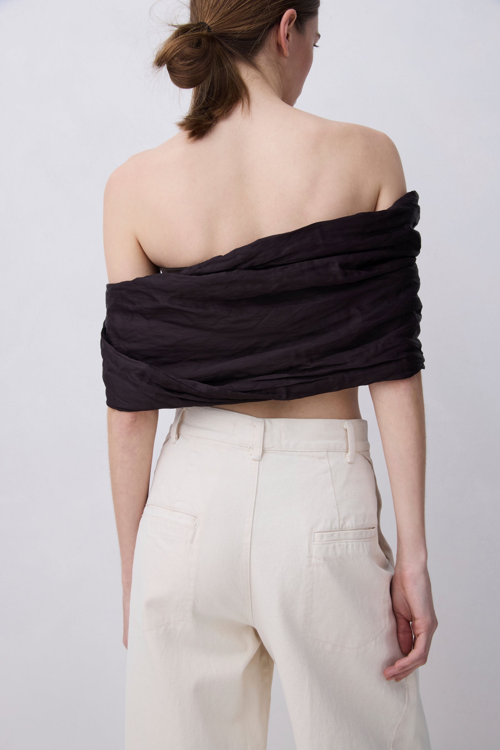 A woman shown from the back wearing a black off-the-shoulder ruched crop top and cream-colored wide-leg trousers, set against a plain light gray studio background.