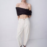 A model stands against a light gray studio background wearing a black off-the-shoulder wrap top, cream wide-leg trousers, and black strappy sandals.