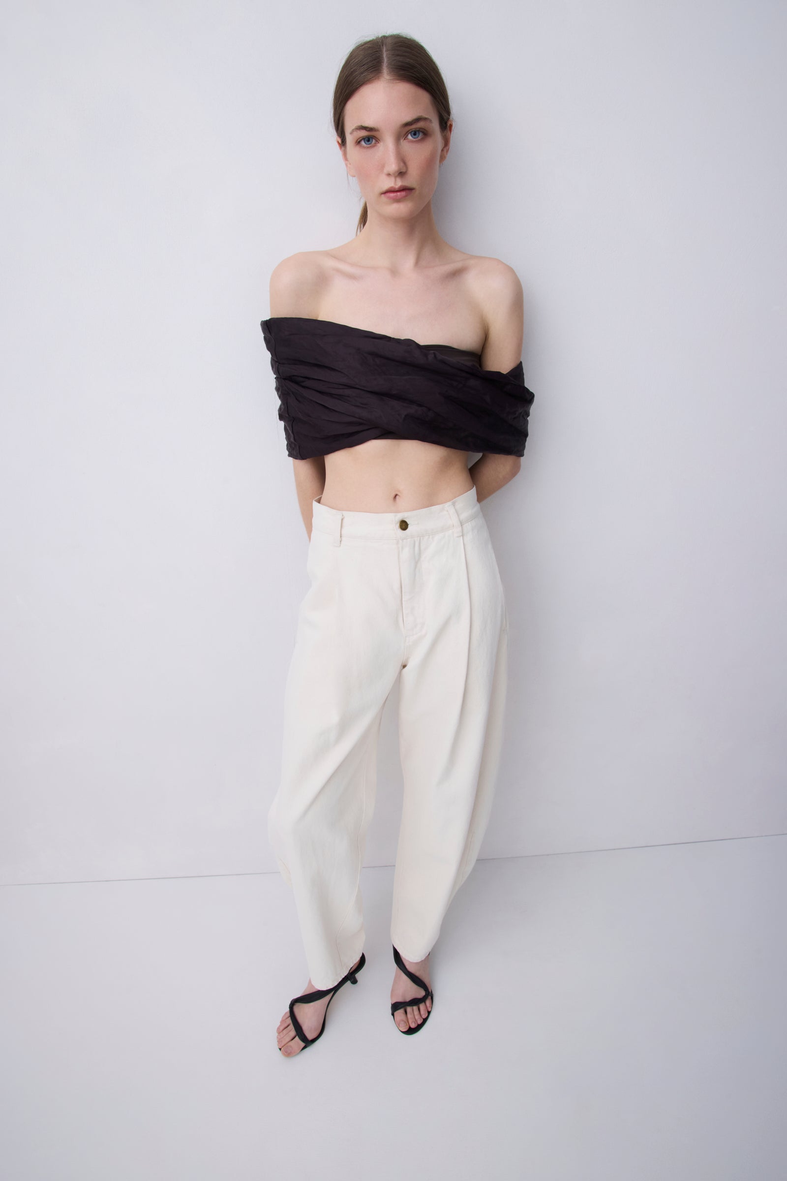 A model stands against a light gray studio background wearing a black off-the-shoulder wrap top, cream wide-leg trousers, and black strappy sandals.