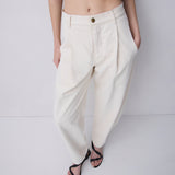 A model wearing off-white pleated wide-leg trousers with a single gold button, styled with a black cropped top and black strappy sandals against a light gray studio background.