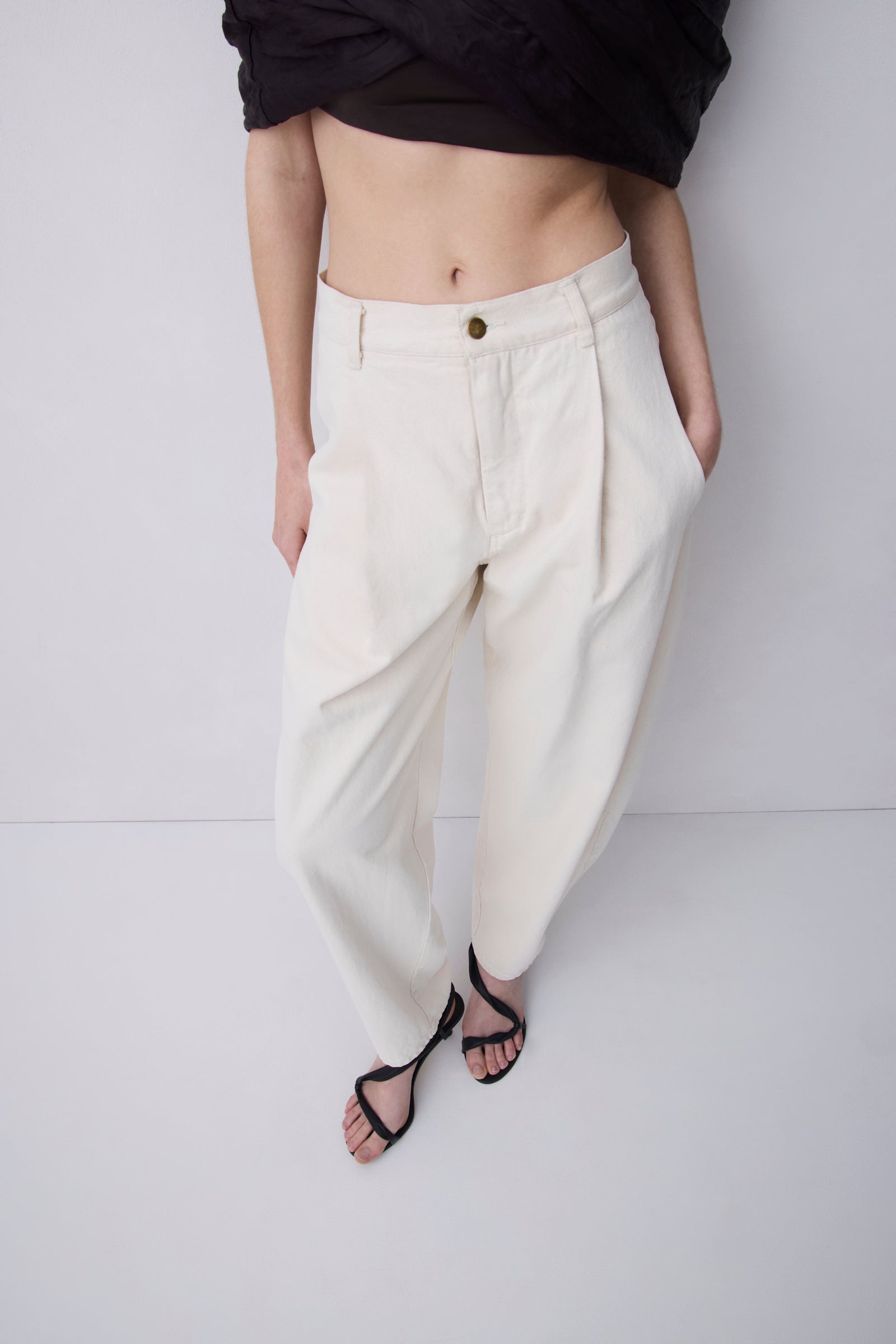 A model wearing off-white pleated wide-leg trousers with a single gold button, styled with a black cropped top and black strappy sandals against a light gray studio background.