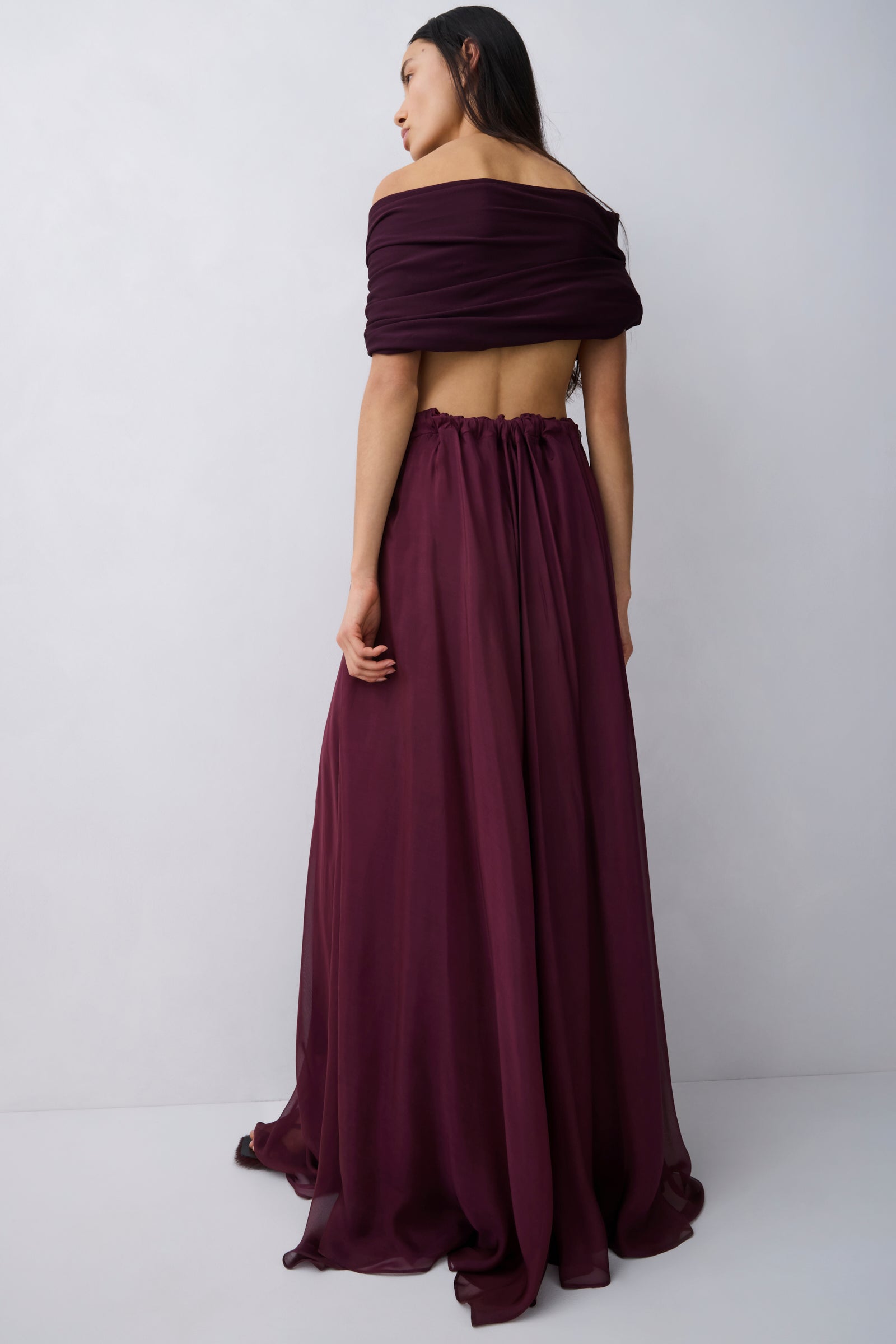 A woman wearing a deep burgundy off-the-shoulder maxi dress with a flowing skirt, shown from the back against a light gray studio background.
