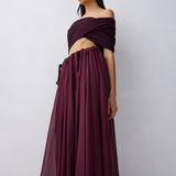 A woman wearing a deep burgundy off-the-shoulder maxi dress with a flowing skirt, shown from the back against a light gray studio background.